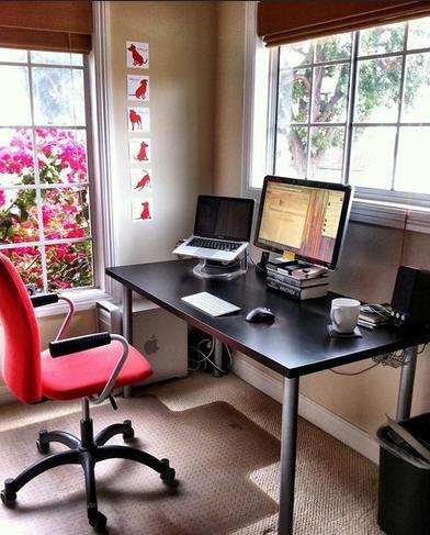 Home office with red office chair, black table, monitor and laptop on stand.