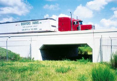 Red and white truck crossing a white bridge with a blue sky background, grassy foreground